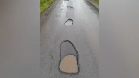 Wilson Beggs Potholes on a wet road. Brown water is sitting in them.