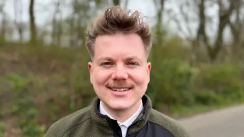 A man with a moustache and light brown hair, Nathan Goldup-John, is smiling.