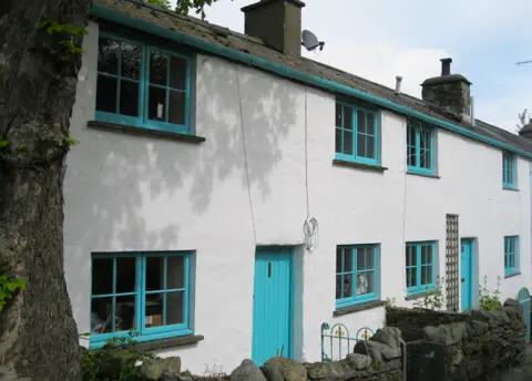 Alan Fryer/Geograph Whitewashed terraced cottages with turquoise doors and cross-hatched traditional window frames. There is a tree trunk to the left of the photo. Dry stone walls border each house and the wrought iron gates are painted the same turquoise colour.