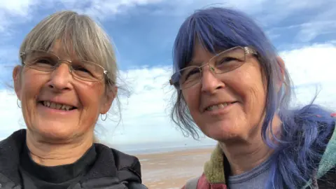 Wendy Ronaldson Two women with silver framed glasses smile at the seaside. The woman on the left has grey hair, silver glasses, and wears a black coat and black jumper. The woman on the right has blue hair, silver glasses and wears a patchwork coat with a blue t-shirt. Behind them is a beach, with the sea far out and a blue sky dotted with clouds above them.