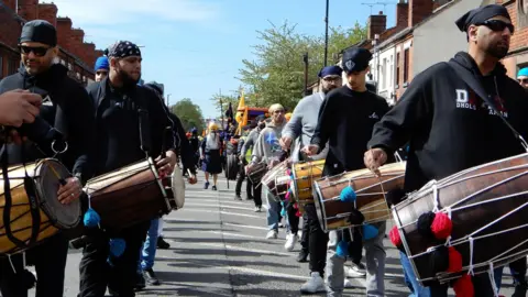 Gurmit Sandhu and Ranjit Sahota Men lined up in a parade with long drums on straps around their neck. The drums are decorated with red blue and black pom poms. The men nearest the camera are wearing black head coverings such as bandanas, turbans and patkas, and dark jumpers. Others can be seen behind them in a procession, and a large vehicle preceding a float.