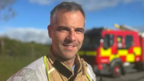 NIFRS Group Commander Andy McVeigh is smiling at the camera. He is standing in front of a red and yellow fire truck. He has grey hair and is wearing a yellow fire-proof suit. 
