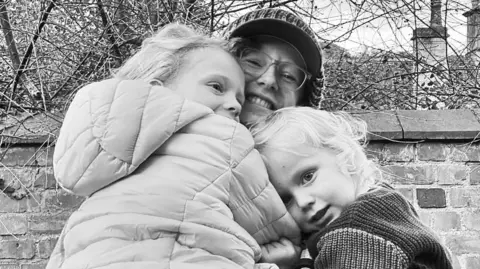 Family handout A black and white image of two young children with light hair being picked up by their mother, who is smiling. They are cuddled in to their mum and are dressed in coats and jumpers. Their mother is wearing a hat and clear rimmed glasses. They are stood in front of a brick wall and a bare tree.
