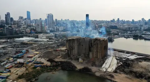 Getty Images Remains of a storage building in Beirut which was destroyed in a large explosion. The severely damaged structure is surrounded by port facilities and shipping containers. The city of Beirut is in the background.