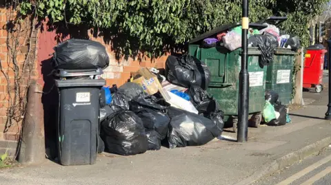 Tempat sampah roda yang meluap, kantong sampah di lantai dan tempat sampah industri yang meluap dengan tas nongkrong