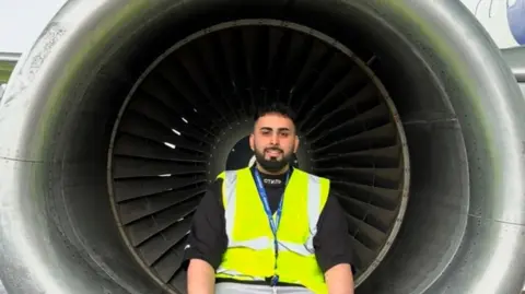 Faizan Mujtaba Faizan Mujtaba, with a beard, is smiling. He is sitting in the centre of the photo wearing a high-vis jacket and a lanyard.