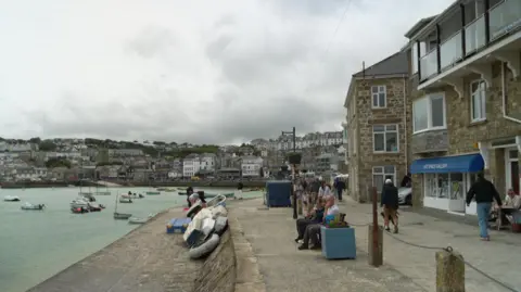 A harbour scene shows a stone promenade lined with buildings, with small boats floating on calm water and a town spread across the hillside in the background under a cloudy sky. People are sitting and walking along the waterfront near stacked dinghies, while a gallery and other seaside shops face the quay.