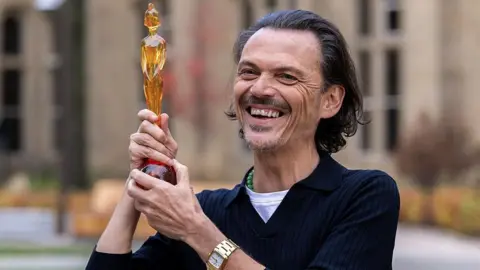 Matthew Williamson, with mid-length dark hair and a goatee beard, wears a dark blue jumper and white T-shirt and holds aloft his Brit Awards design, a yellow-amber statuette of a woman wearing a crested helmet, outside the brick-built walls of the University of Manchester