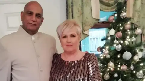 Bobby Rao Bobby Rao is standing beside his wife Josephine. There is a Christmas tree covered in baubles and light beside her. She has short blond hair and is wearing a glittery gold top. He is wearing a beige formal jacket/shirt which has no button at the top. 
