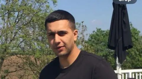 Family photo Kamran Aman, wearing a black t-shirt, standing in front of a number of trees