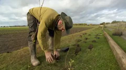 A man, with a beard and wearing a heat, wellies and green clothes, planting trees as part of the Humber Forest