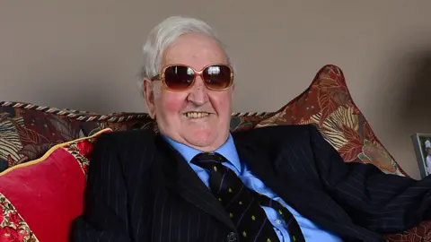 Pacemaker Sir Kenneth Bloomfield has grey hair and is wearing brown sunglasses. He is sitting on a red and brown sofa and is smiling at the camera. He has a navy suit on with a blue shirt and striped tie.