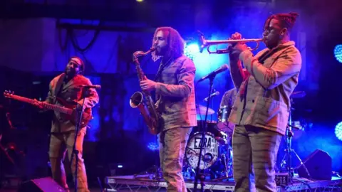 Three members of the band Ezra Collective perform on a stage. They are three men in matching brown suits. One man plays a bass guitar, one plays a saxophone and another plays a trumpet.