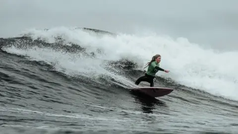 Oscar James The picture shows a person riding a wave on a surfboard in the ocean. The surfer is wearing a green top and black wetsuit, leaning forward with one arm extended. The wave behind them is large and foamy, indicating strong surf conditions. The water is dark and choppy, and the sky looks overcast.