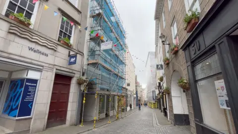 The image shows a narrow cobblestone shopping street with colourful bunting overhead. On the left, a Boots store is next to a building wrapped in scaffolding and blue netting. On the right, buildings with flower boxes line the pavement. Hanging shop signs, including “Accessorize,” are visible down the street, which is pedestrianised and busy with renovation work.