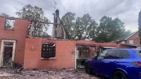 A building with a missing roof and a lot of fire debris on the outside. A dark blue car in good condition is parked outside it.