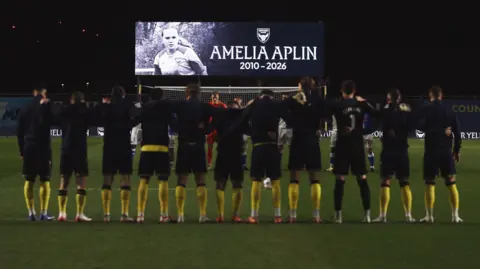 The eleven Oxford United team players on the pitch facing a screen with a tribute to Amelia Alpin above the net. They are wearing black kits with yellow socks.
