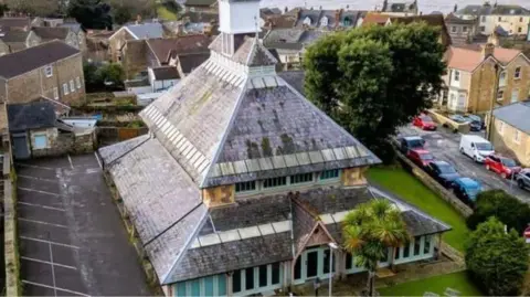 A large building with a tall sloped roof and spire covered in moss.