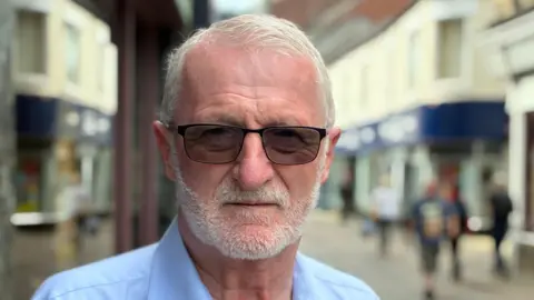 Don Threadgold. Close up of his face with sunglasses on. He is wearing a pale blue shirt. A town centre can be seen in the background. It is blurry. 