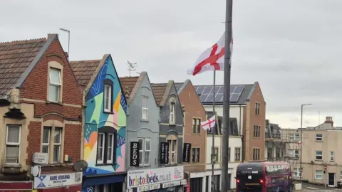 Two St George flags can be seen flying on lampposts. The high street is Duckmoor Road in Bedminster, where there is a row of shops and a bus on the road.