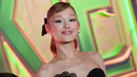 Getty Images Ariana Grande smiling close up to the camera, she is wearing a black strapless dress. She has her light brown hair tied up in a ponytail with a black bow, she has a short side fringe. Behind her is the Wicked logo in huge print. 