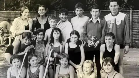 Children at the lido after a swimming gala. The black and white photograph shows boys and girls lined up, some in swimming costumes, near the steps to the pool. Some have towels around them. One boy is holding a cup after the competition.