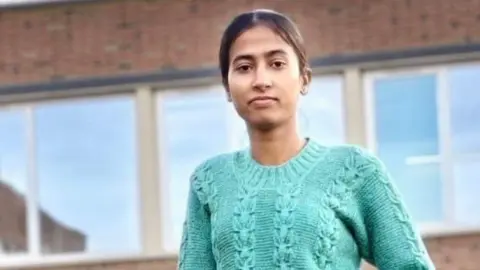 Family handout A woman in a teal knitted jumper and dark trousers stands outside in front of a red brick building. She has brown hair that has been tied back.