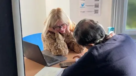 Legal Aid Woman with blonde hair and red glasses sitting at a desk by a laptop looking at a phone being pointed to by a man with grey hair, who is wearing a black jumper. 