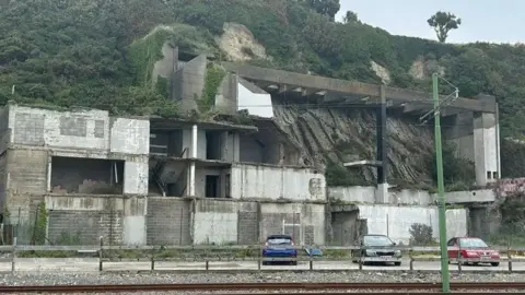The outside of the site, you can see the remainder of a derelict building on the cliff face, there are cars in front.