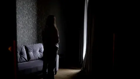 A person in shade with their back to the camera stands by a sofa 