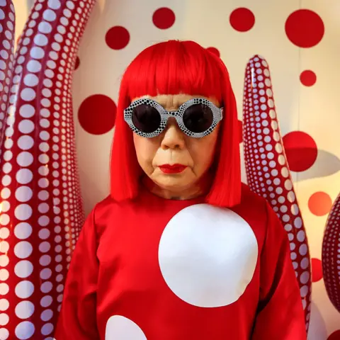 Gamma-Rapho via Getty Images Yayoi Kusama looks at the camera wearing round glasses and a red top with white polka against a backdrop of polka dot tentacles for her exhibition in Hong Kong in 2012.
