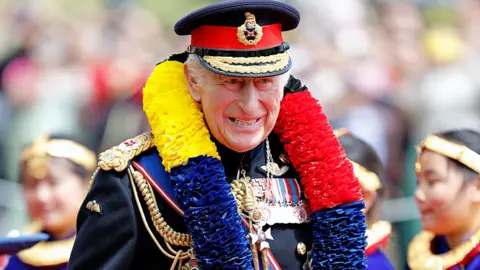 Max Mumby/Getty Images King Charles in ceremonial uniform and a row of medals and wearing a huge boa in red, yellow and blue colours. He is smiling to camera and there are people at the Ghurkha event blurred in the background