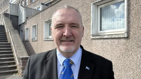 A middle-aged man wearing a blue shirt and a black suit with a bright blue tie. He has a goatee and is smiling at the camera. He is standing in front of a hospital building with windows. 