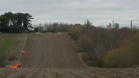 Jay Naylor Films Volunteers in orange high vis walking along an undulating field towards the site of a new grove being planted, with the Ketton Cement Works visible on the horizon