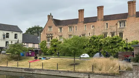 Magdalene College, Cambridge, with a grass area in front with a handful of protesters on blankets an a couple of round tents