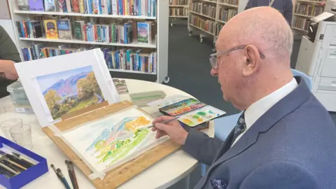 Laura Coffey/BBC A man wearing glasses, a tie and a blue suit with a handkerchief in the jacket pocket and white shirt. He is sitting at a table doing a watercolour painting. He has his paints to his right and a box of paint brushes to his left.
