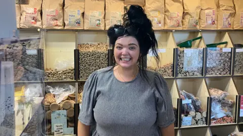 A woman wearing a black and white gingham dress stands behind the counter at a pet food store. She has long black hair in a big top knot and sunglasses on her head. She is smiling. There are containers with dog treats and scoops behind her.