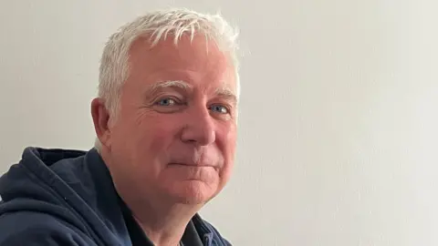 A man with white hair. He is wearing a hooded top.