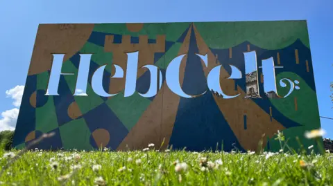 A free standing board advertising HebCelt festival. It's painted brown, green and blue and the name of the festival is cut out in large letters. A sunny day in the background. 