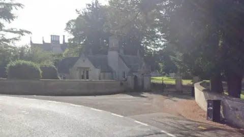 Google A gatehouse with stone walls at the entrance to a country house. The house itself is visible behind the gatehouse and trees.