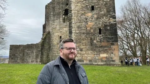 David McDonald - a man standing in front of a castle, with glasses, short dark/greying hair and a grey jacket and black top.