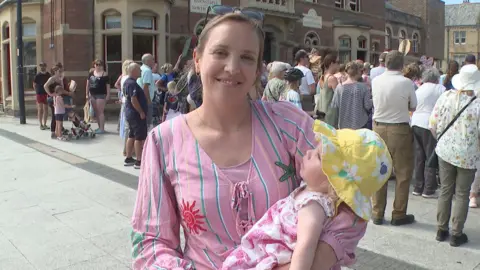 Sarah Henshall wearing a pink striped top holds her baby in a yellow sun hat while standing in a town square. A crowd has gathered outside a large brick building in the background.