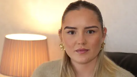 A young woman with long blonde hair smiles at the camera. She has dark eyebrows and a nose ring. She is wearing a light-coloured top and is sitting in front of a pale background.
