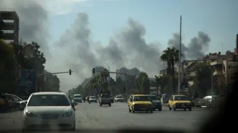 Getty Images Rook stijgt op aan de horizon van een straat in Damascus, Syrië. Er rijden auto's over de weg. Het is dag met een blauwe lucht.