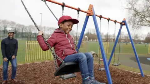 Boy smiling on the swing and his father behind smiling 