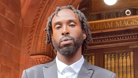 Omar Abdirizak dressed in a black suit and white shirt, stood in front of a red brick and wooden doors of Birmingham Magistrate's Court