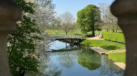 EstherJ A white footbridge crosses a river. Trimmed lawns and hedges are also in shot.