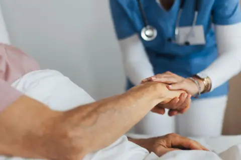 A doctor or nurse is holding the hand of someone lying in a hospital bed. You cannot see anyone's faces.