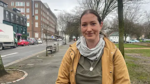 White woman in grey scarf and silver necklace looks into the camera. she stands by a park next to a main road in Jersey.