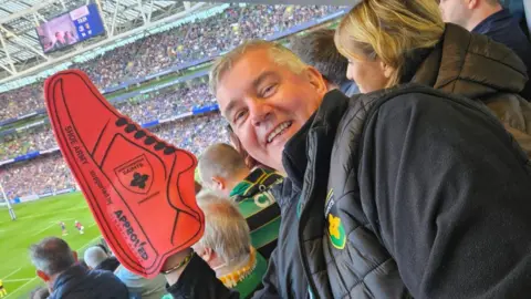 Colin Wells Colin Wells in a Northampton Saints jacket with a red foam shoe over his hand. He is standing against the backdrop of the Aviva Stadium in Dublin during the match against Leinster.